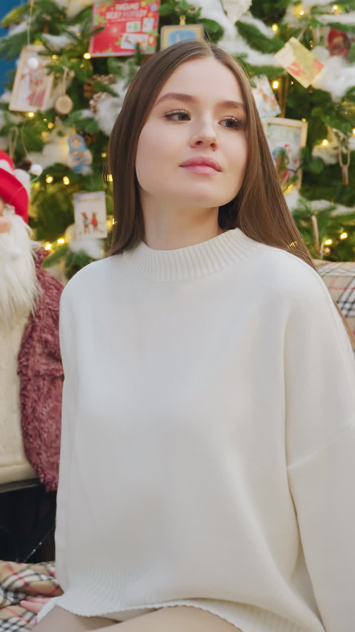 Young woman in brown trousers and white top walks up and sits on chair, relaxing in festive church decorated with Christmas decor, including trees, lights, and reindeer figures