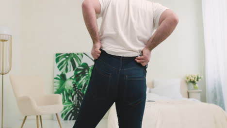 rear view of man buttoning tight jeans with difficulty in the bedroom