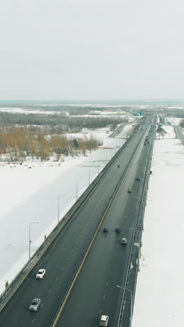 cars drive along huge long gray bridge making shadow on white frozen river against dense forest upper view