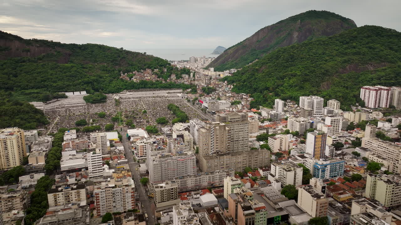 Botafogo, Rio de Janeiro, contrast between dense cityscape and sprawling São João Batista cemetery at the base of lush green mountains, Brazil. Aerial forward