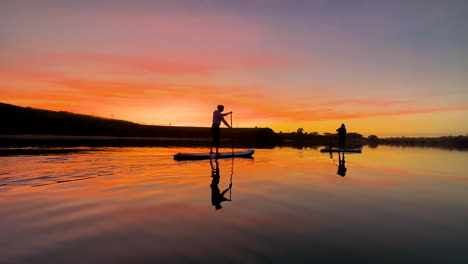 Zwei-Silhouetten-Von-Ein-Paar-Mädchen,-Die-Einen-Wunderschönen-Sonnenuntergang-Auf-Dem-Paddleboard-Mit-Einer-Fantastischen-Wasserreflexion-In-Südkalifornien-An-Einer-örtlichen-Lagune-In-Carlsbad,-Ca