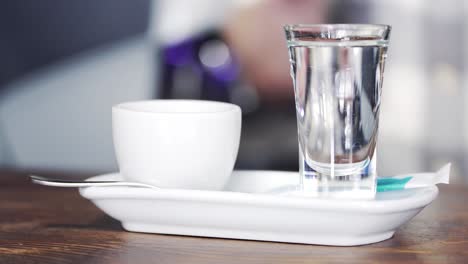 barman puts a cup of coffee, teaspoon, some sugar and a glass of water on the wooden counter. close-up