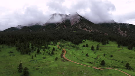 Brown-dirt-trail-with-hikers-snakes-through-green-meadow-toward-Chautauqua-Park-pine-forest-and-misty-Flatirons,-aerial-dolly,-Boulder-Colorado