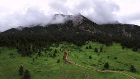 Brauner-Feldweg-Mit-Wanderern-Schlängelt-Sich-Durch-Grüne-Wiese-In-Richtung-Kiefernwald-Im-Chautauqua-Park-Und-Nebligen-Flatirons,-Luftwagen,-Felsbrocken-Colorado