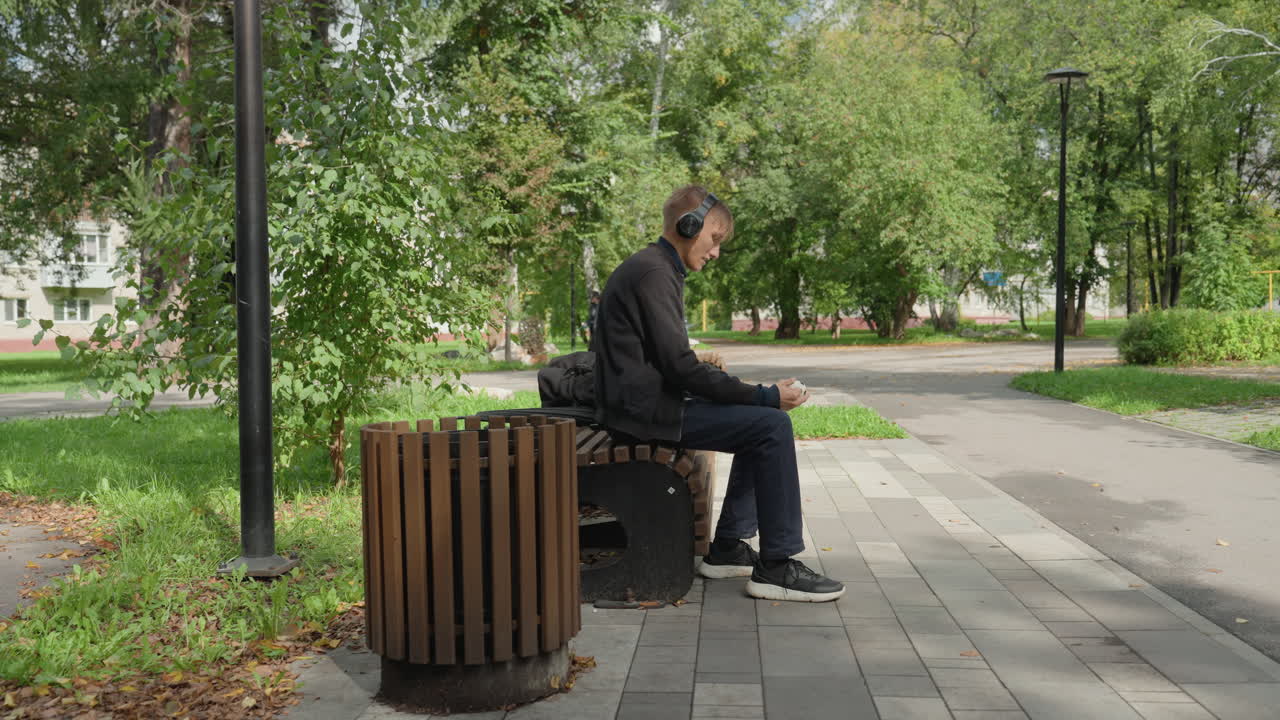 Uomo seduto su una panchina del parco che ascolta la musica con le cuffie, scorrendo il telefono, cestino di legno accanto alla panchina, sentiero lastricato, alberi frondosi, cielo nuvoloso, umore pensieroso, spazio verde urbano, momento di tranquillità