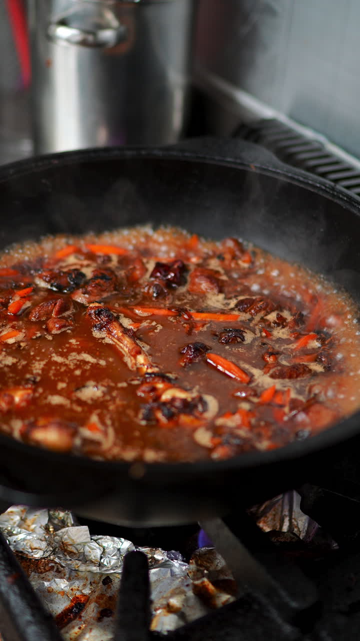 Professional chef reducing sauce with meat and vegetables in heavy duty pan, simmering over intense gas flame inside commercial kitchen environment