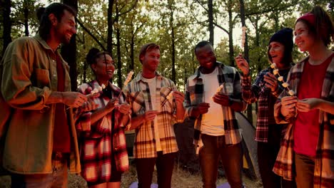 A-happy-company-of-six-people-in-special-camping-clothes-in-plaid-shirts-stand-around-a-fire-and-string-marshmallows-onto-small-wooden-sticks-in-a-green-autumn-forest.-The-fire-illuminates-the-hikers-with-red-orange-color-during-the-evening