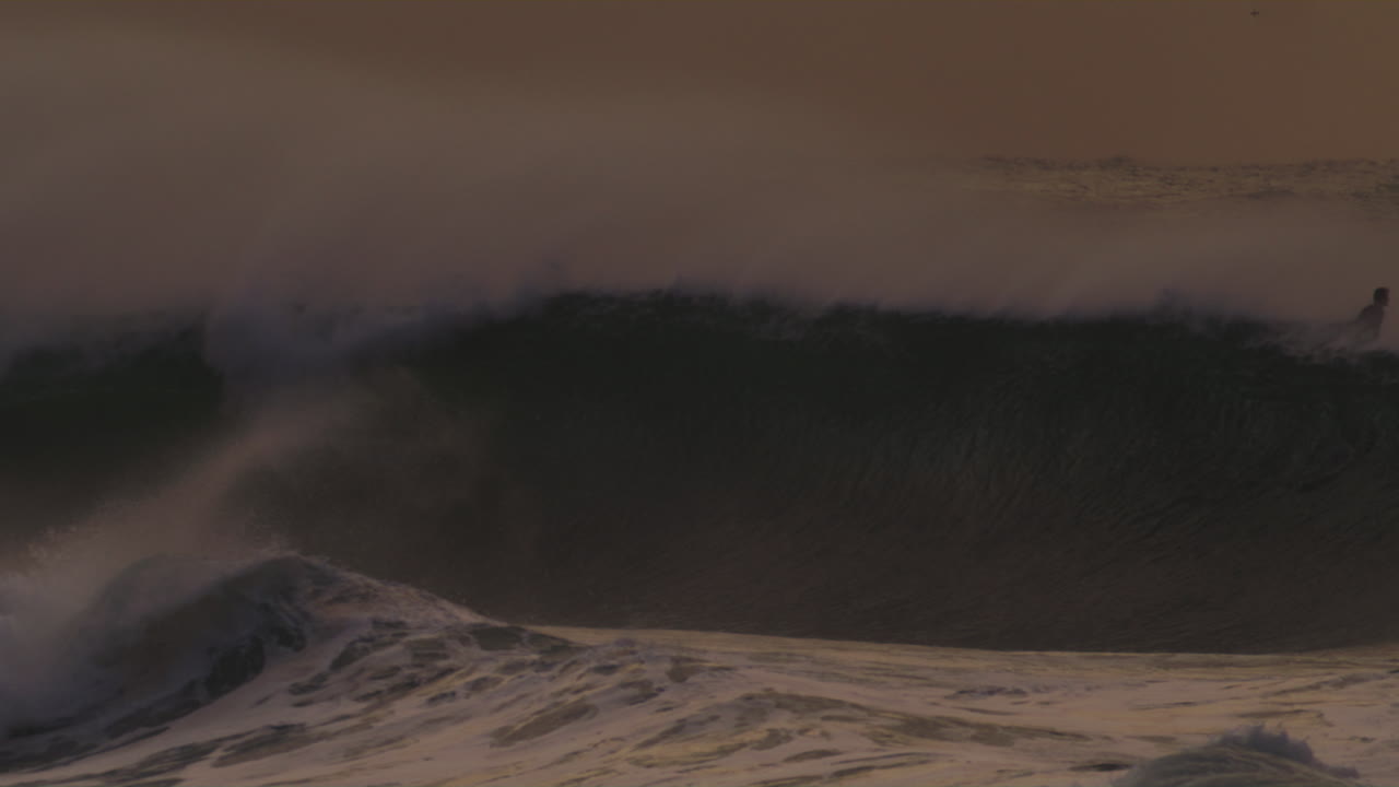A striking slow-motion capture of a sunset wave crashing powerfully, filmed from the shore, highlighting the wave's dark silhouette and evening light