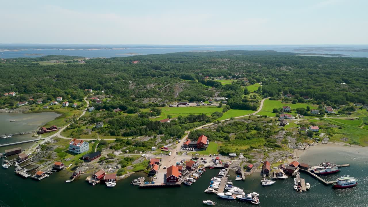 Drönare drar sig tillbaka för att avslöja fiskebyn på Sydkoster med röda båthus, färjekaj, förtöjda båtar och frodigt grönt landskap omgiven av den svenska skärgården.