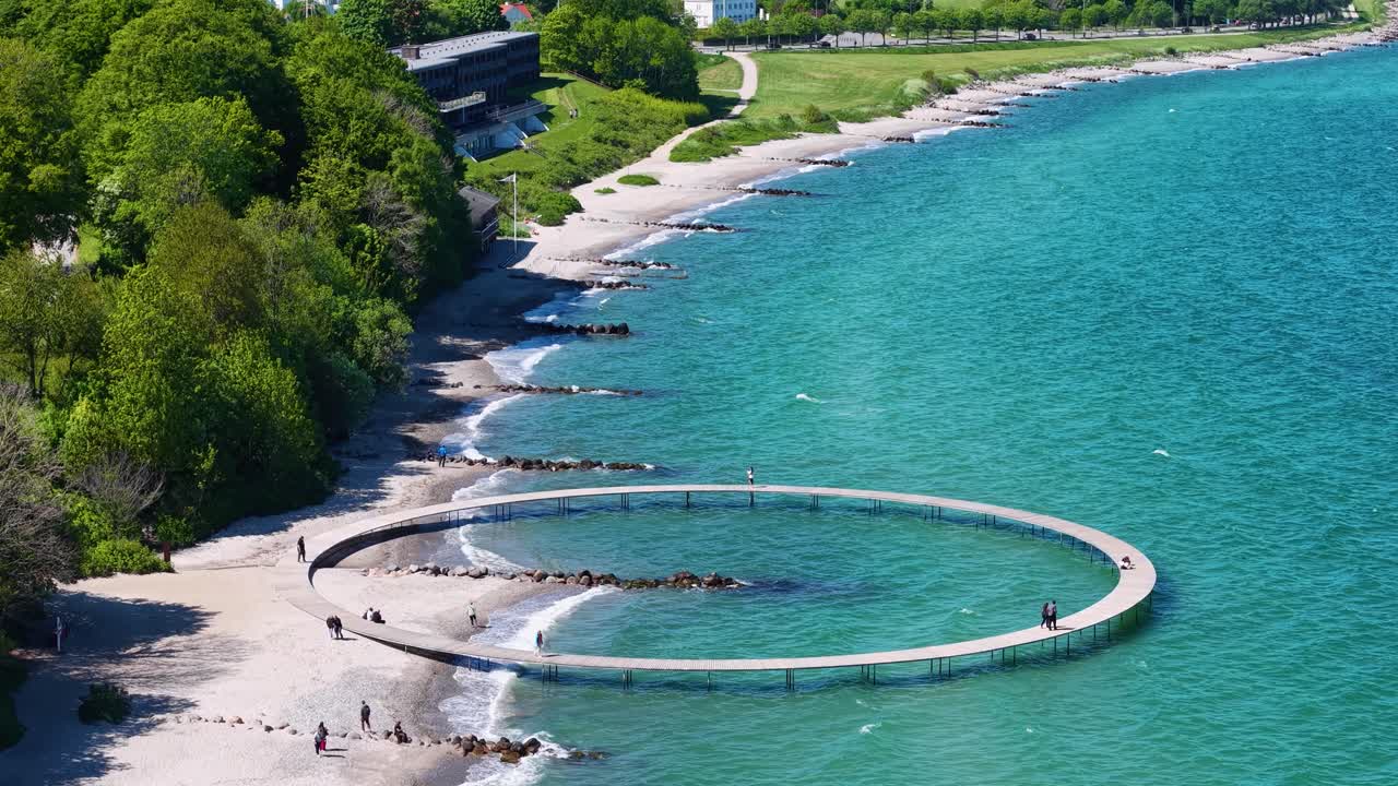 Aerial view of Denmark’s Infinite Bridge curving over turquoise water and white sand beach