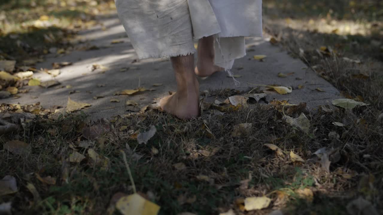A dramatic slow motion closeup of the feet of a middle eastern man dressed as Jesus Christ walking down a sidewalk while he wears a white, tattered robe