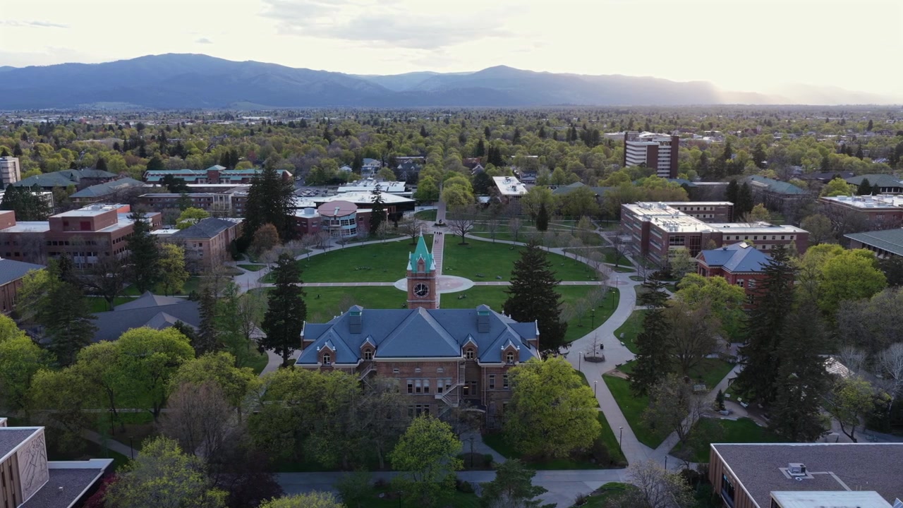 Storico campus dell'Università del Montana a Missoula con prato verde centrale, edifici accademici e montagne circostanti che brillano nella calda luce dell'alba primaverile. Atmosfera scenica calma. Ampia ripresa aerea