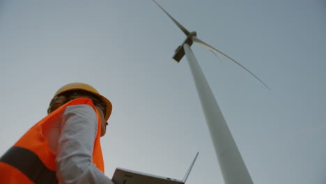 lower view of a caucasian female engineer wearing a helmet and using laptop under the huge windmill turbine and watching it working and producing energy