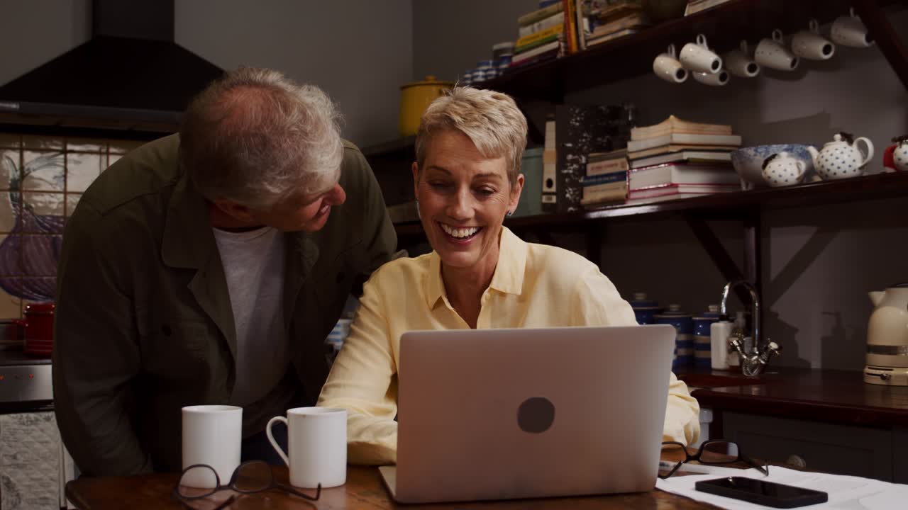 una pareja de mayores caucásicos se ríen en la cocina mientras escriben en la computadora portátil.