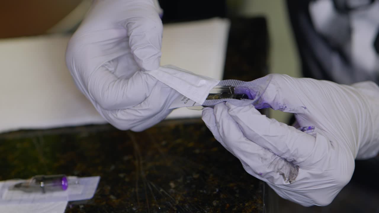 artista del tatuaje con guantes y tinta en todas las manos abriendo una nueva punta de aguja para la máquina rotativa, sacándola del paquete y colocándola