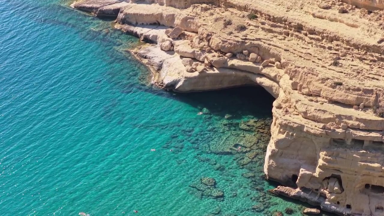 Vista aerea con drone di una scogliera di arenaria chiara e un'ampia grotta marina sopra acqua limpida e turchese, Matala, Grecia