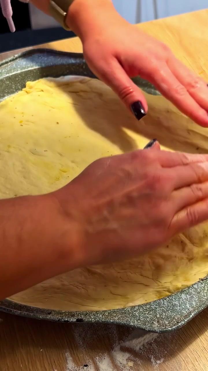 Hands gently pressing and stretching pizza dough evenly into a round pan on a wooden surface, preparing for baking.
