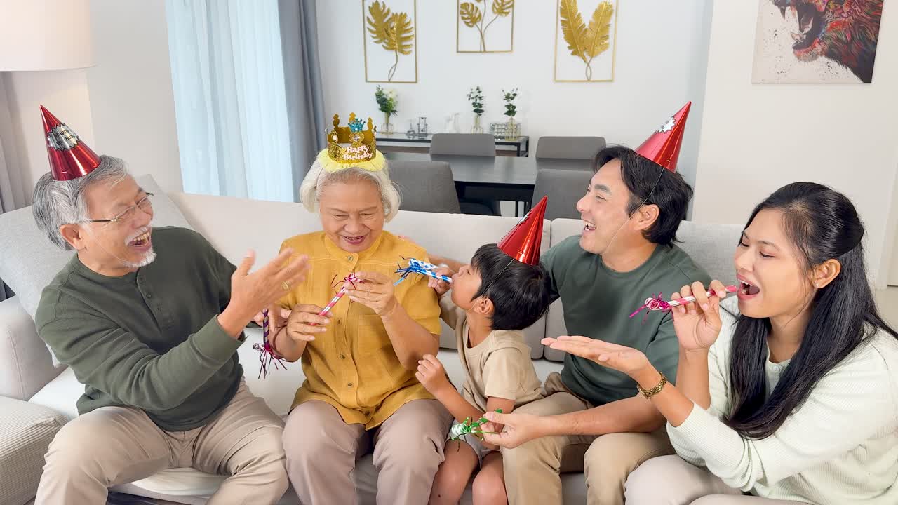 Four Asian family members celebrate with party hats and blowers, laughing together in bright living room