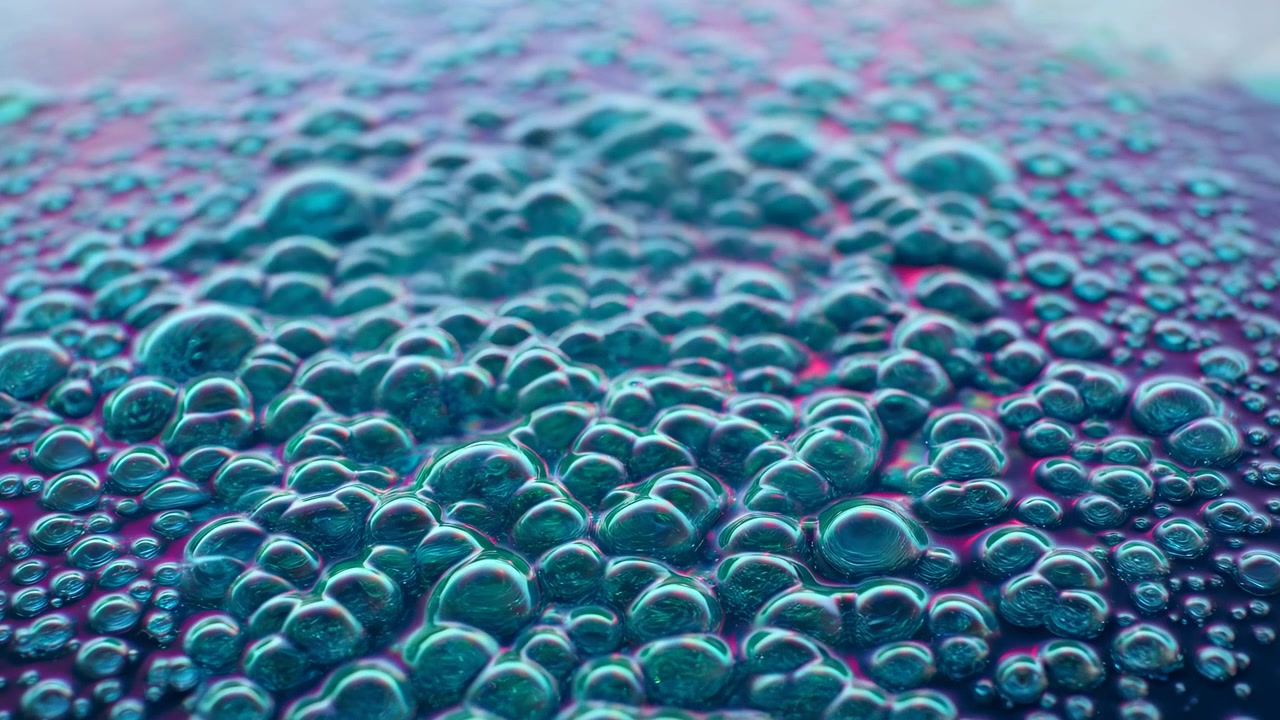 Macro primo piano di bolle di sapone raggruppate e schiuma su una superficie piana lucida, illuminate con illuminazione da studio iridescente color verde acqua e viola. Bolle arrotondate di varie