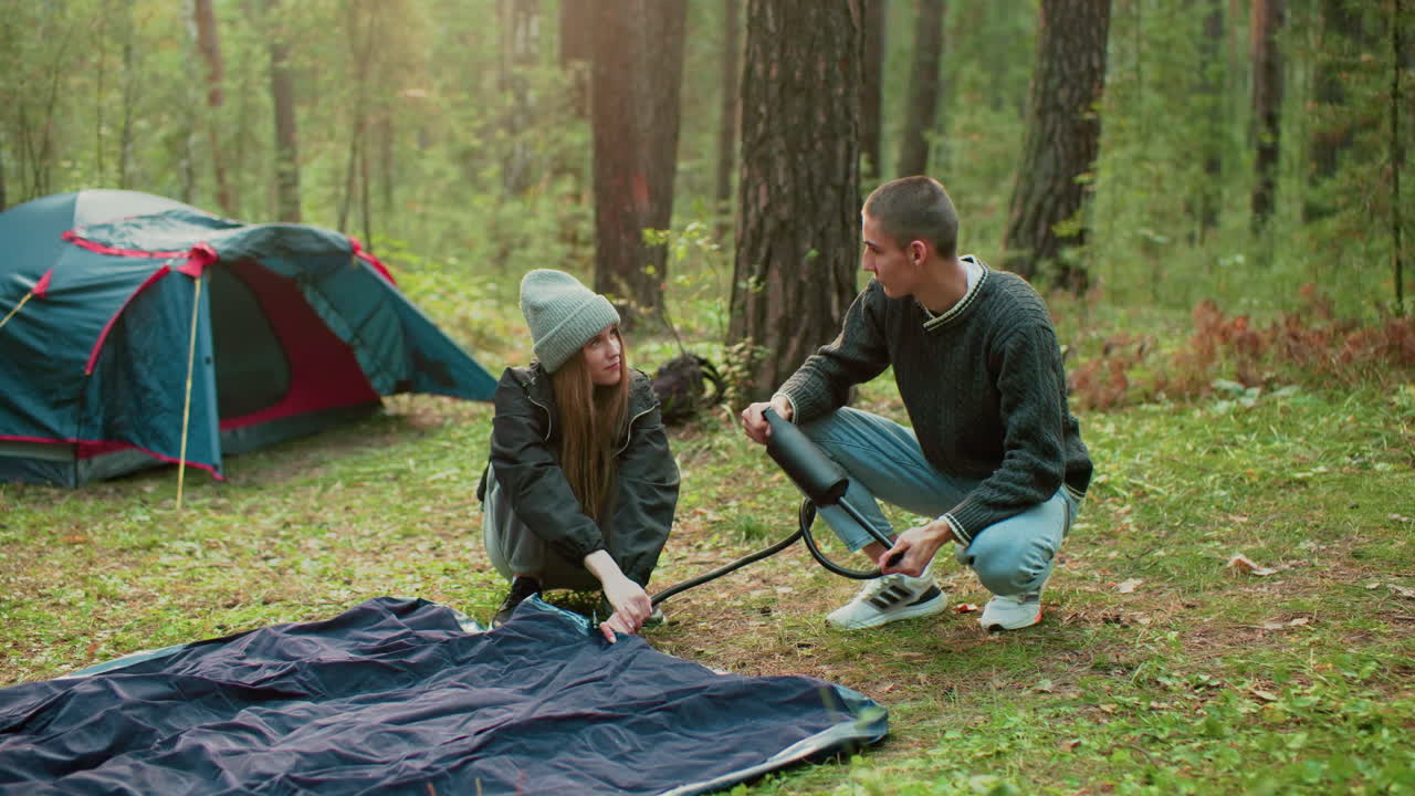 Woman gazes at her partner while checking tent bag as man pumps air into it, surrounded by forest trees and pitched tent, showing teamwork and bonding during outdoor camping adventure