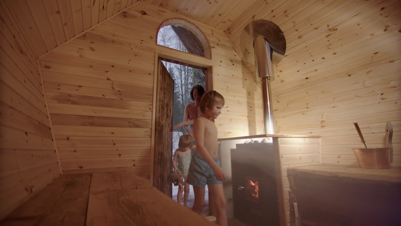 Mom and two kids enters wooden sauna then ladles water on rocks of wood stove