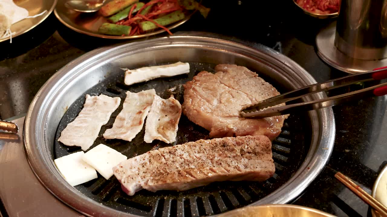 Hand uses metal tongs to flip pork slice on tabletop Korean barbecue grill, warm lighting