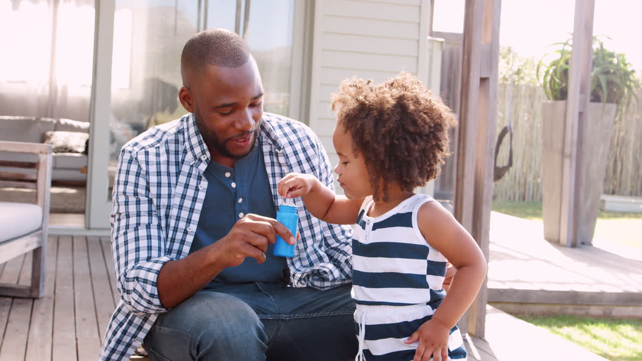 Young black girl blowing bubbles with dad outside house