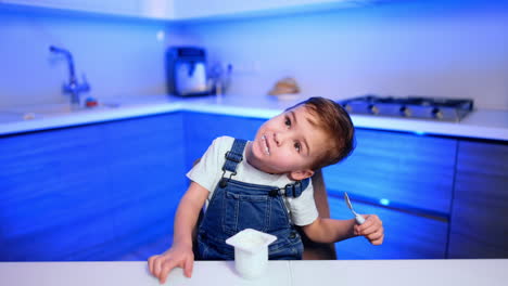 lovely baby boy wearing white t-shirt and jeans romper sits at table in the kitchen. kid eats yogurt and waves his head.
