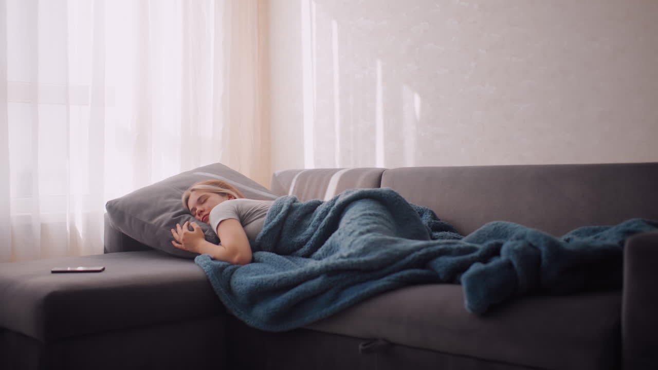 Fatigue lady lying on couch holding blanket trying not to wake up phone near by as sunlight streams softly through curtains calm peaceful indoor atmosphere reflecting comfort