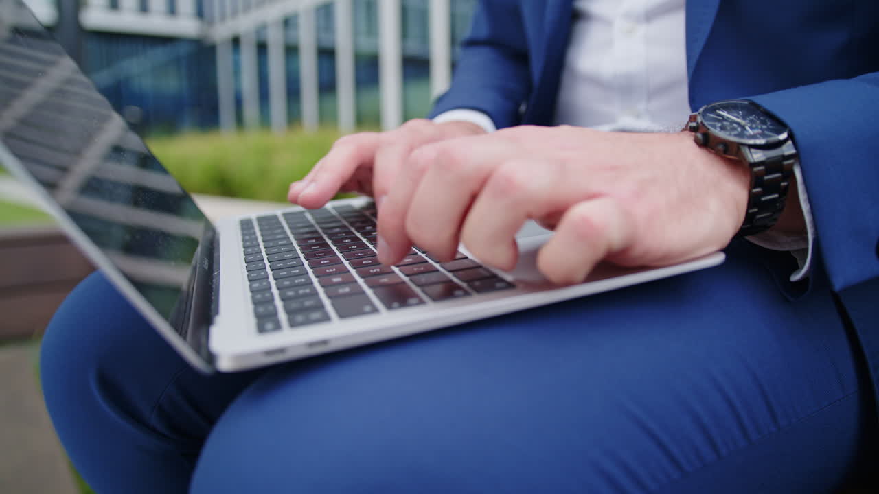 A businessman in a blue suit sits with a laptop in front of the office building