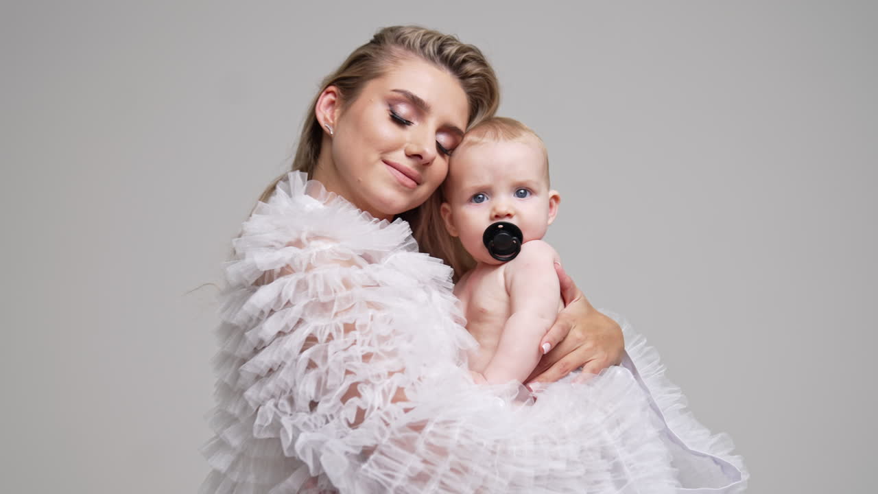 Caucasian mother with her lovely baby with pacifier in mouth. Woman waves her child looking at camera. White backdrop.