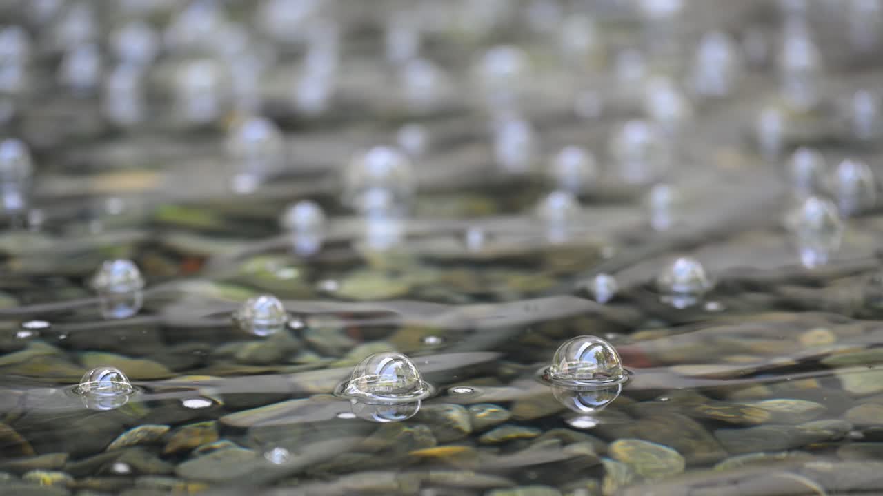 Heavy rain or a small waterfall splashes into clear, shallow water, creating an effervescent scene. This close-up, abstract shot captures the resulting air bubbles as they form on the surface