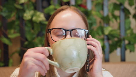 close up cheerful woman wearing glasses smiles while talking on phone during breakfast in cozy cafe with green plant decor in background enjoying relaxed morning conversation indoors
