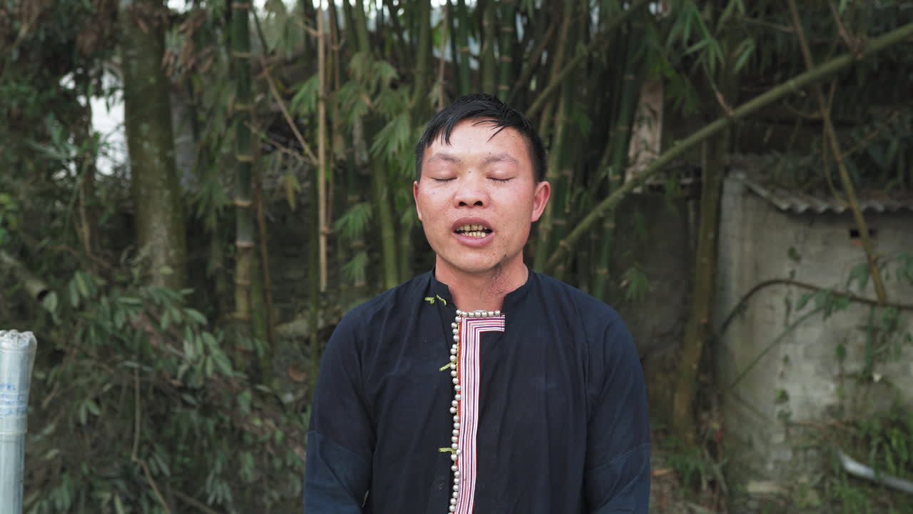 A young Black Dao man speaks in close-up. He wears traditional dress. Vegetation and a small shelter appear behind him.