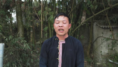 a young black dao man speaks in close-up. he wears traditional dress. vegetation and a small shelter appear behind him.