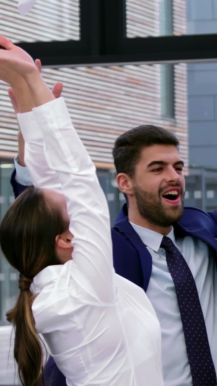 Video verticale: Colleghi in abito e camicetta che festeggiano lanciando carte dopo l'approvazione, visibili dalle finestre dell'ufficio.