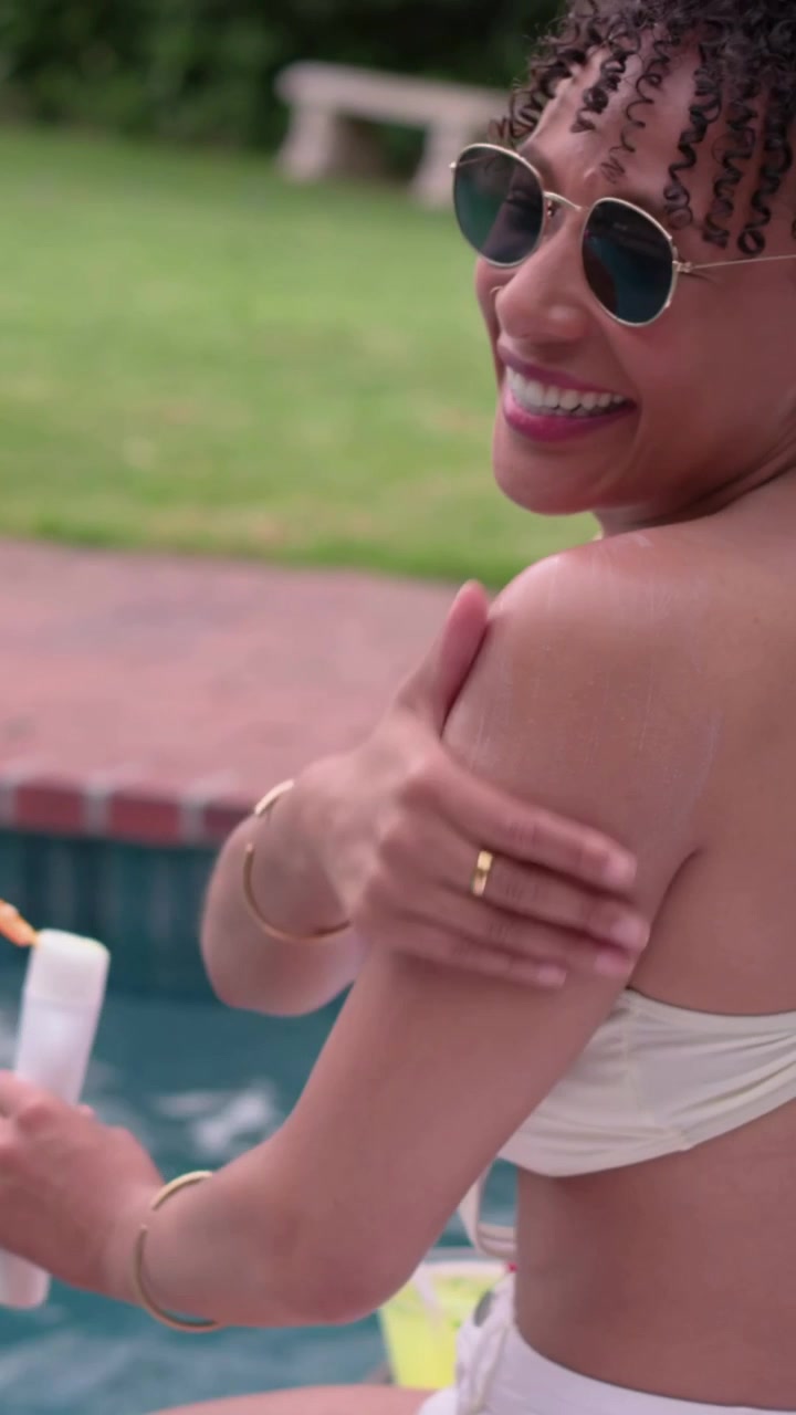 Vertical video: Sun warming woman in white bikini and shorts rubbing arms at pool, holding popsicle