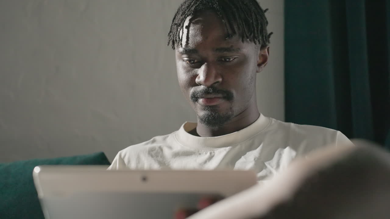 Black youth focused on tablet, seated on teal sofa in modern, peaceful living room. Soft sunlight illuminates his face, creating a calm and thoughtful moment in cozy, minimalist space