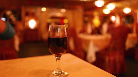 A person’s hand moves and arranges three red wine glasses on a wooden table in a warmly lit, elegant restaurant with a blurred background