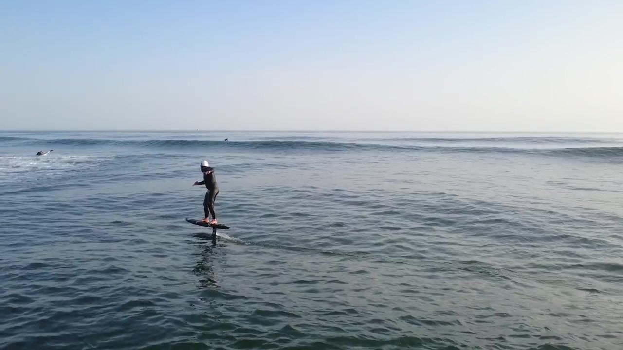 Vista drone di un surfista con foil che "pompa" sull'oceano, alla ricerca di un'altra onda da cavalcare con velocità, fluidità, equilibrio e controllo fluido.