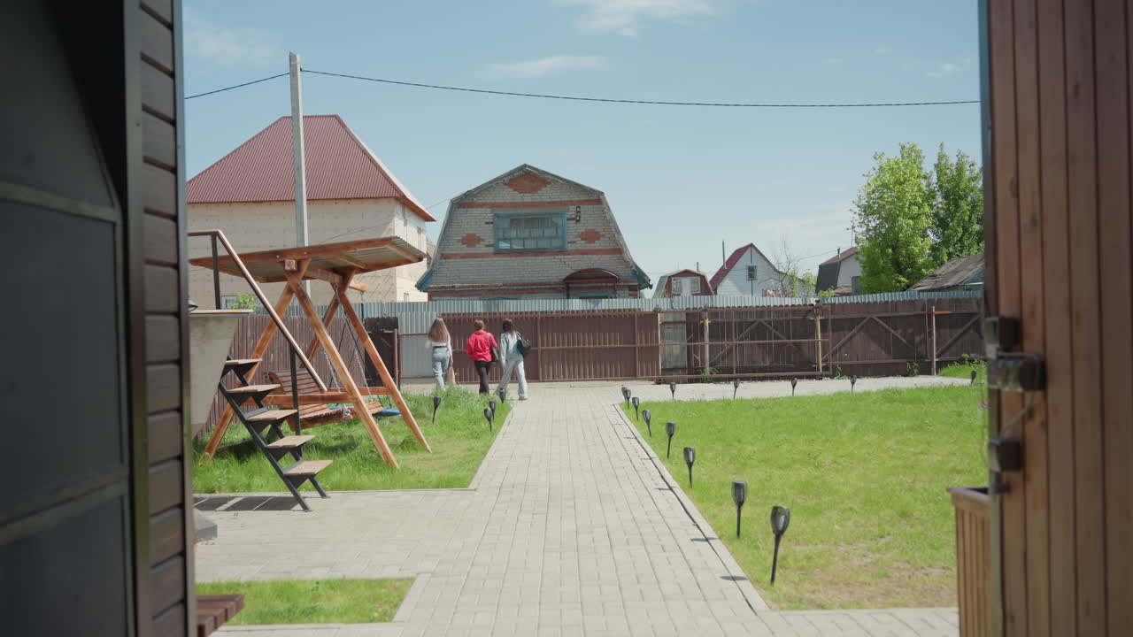 Rear view of two ladies leaving yard, strolling along paved walkway between lawns, passing wooden swing toward brown gate under bright sky in suburban neighborhood