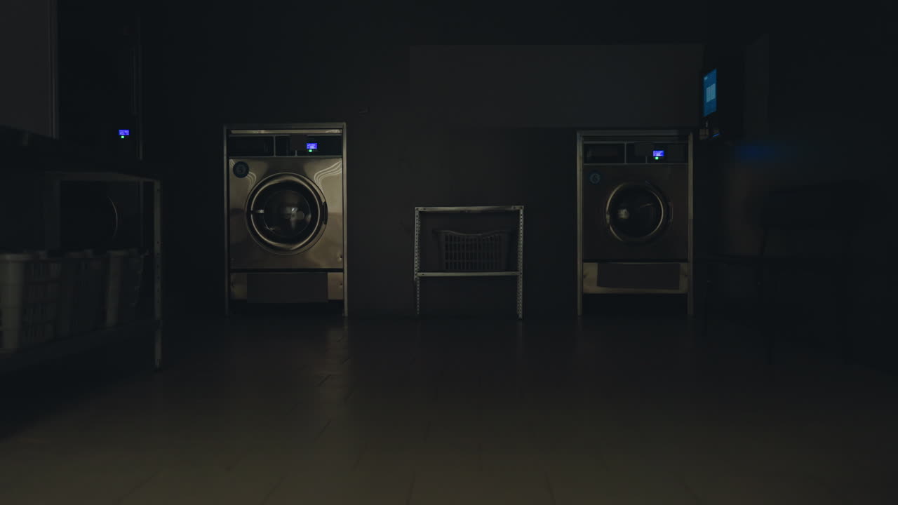 Modern laundry shop interior illuminated after lights turn on, showcasing washing machines, laundry baskets, metal shelf, black bench, and tiled floor in clean service facility environment