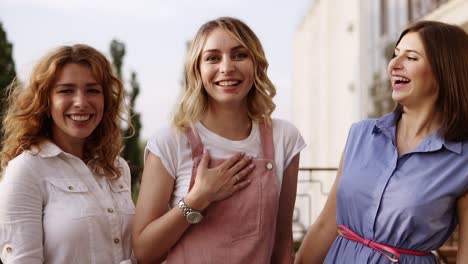 the blonde girl is standing in the middle with excitement and her girlfriends kiss her on the cheeks. hen party. stylish women. outside on a balcony. front footage