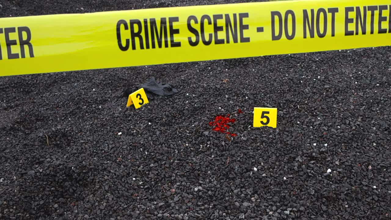 Police yellow colored crime scene forensic tape ribbon in front of yellow markers that are next to a blood stain and a dark blakc nylon glove on dark gravel rocky ground at day time, Shallow depth