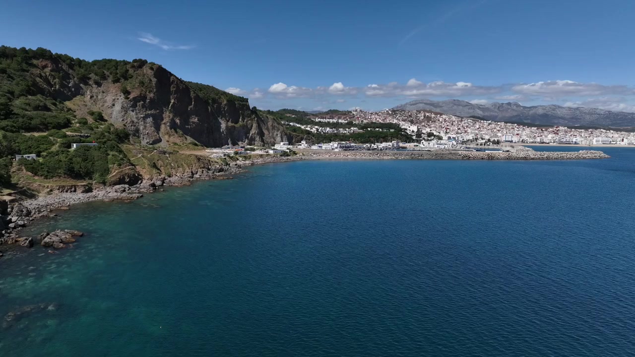 Ampia vista aerea di una costa mediterranea frastagliata nel Marocco settentrionale, con scogliere rocciose, una baia blu calma, una città costiera bianca in lontananza e catene montuose sotto cieli sereni