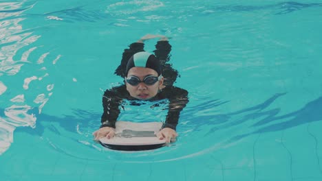 joven asiática usa gorra y gafas de protección aprendiendo a nadar mientras sostiene una tabla de espuma con las piernas trabajando en la piscina