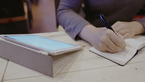 Woman-taking-notes-in-notebook-touch-pad-nearby