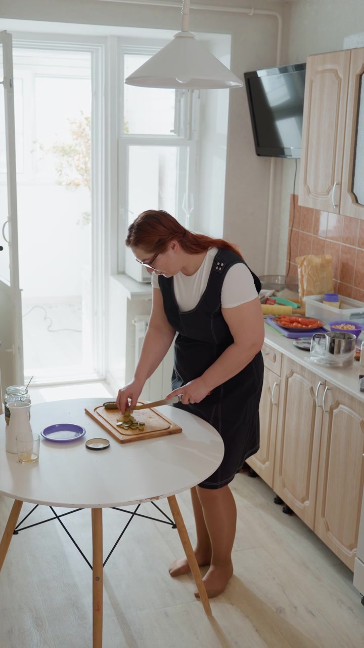 Kvinne står ved et rundt hvitt bord og skjærer brød med kniv mens hun lener seg fremover og arrangerer skiver på skjærebrettet; rolig, fokusert uttrykk og praktisk bevegelse formidler daglig måltidsforberedelse.