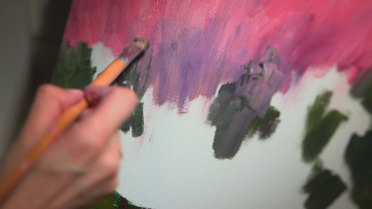 Primo piano di una mano che usa un pennello per applicare vernice verde scuro e nera sotto aree rosa e viola.
