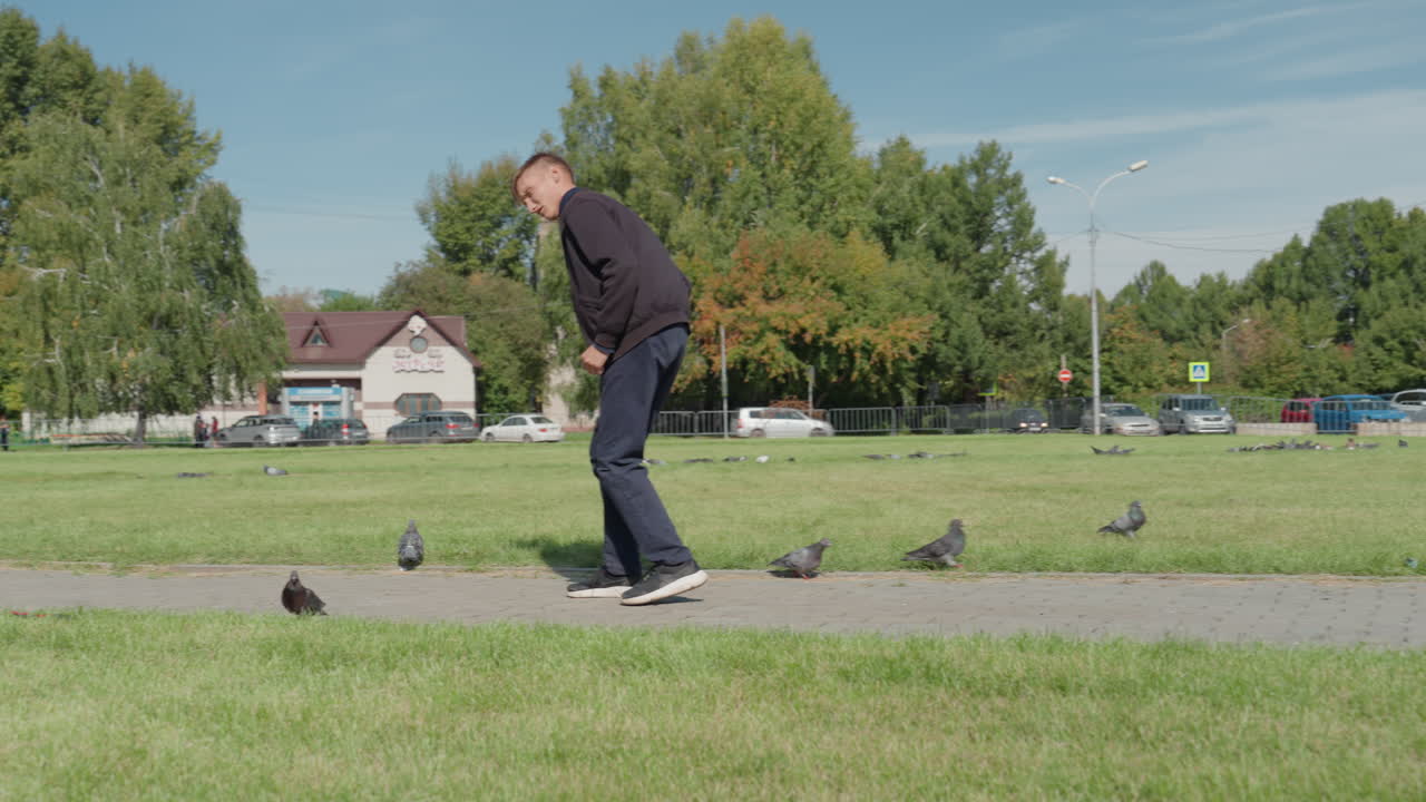 Hvid mand fodrer duer i solrig bypark, sidder på hug på stien og spreder flokken, afslappet jakke og jeans, grøn græsplæne og træer, forstadbygninger i baggrunden, ærligt fritidsøjeblik